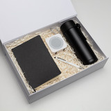 A black notebook, a white speaker, a black drink bottle, and a pen in a gift box with shredded paper.