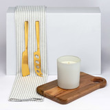 A wooden serving board, a white candle in a frosted glass holder, and golden utensils on a striped cloth. A wooden serving board, a white candle in a frosted glass holder, and golden utensils on a striped cloth.