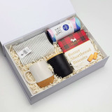A gift box containing a striped cloth, two coffee mugs (one white with a cork base, one black), and shortbread biscuits.