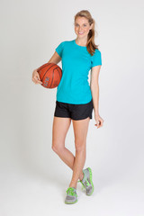 A woman stands holding a basketball, wearing a teal athletic t-shirt and black shorts, with athletic footwear.