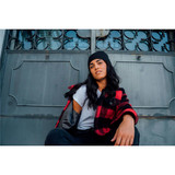 A young woman poses casually, wearing a black and red checkered jacket and a black beanie, against a metallic background.