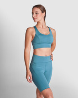 A woman poses in a teal sports cropped tank top and matching high-waisted shorts, featuring a logo.
