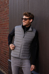 A man wearing a grey down vest stands against a brick wall, smiling and wearing sunglasses. The vest has a logo.