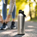 A drink bottle in stainless steel with a black lid, featuring a logo, set on a path surrounded by greenery.