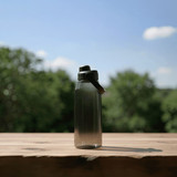 A drink bottle in a gradient of grey with a black lid, placed on a wooden surface against a blurred green background.
