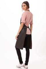 A stylish black bib apron worn over a pink shirt, featured on a model with long hair. Includes a logo.