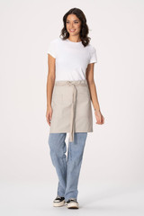 A waist apron in light beige, featuring a tied waistband, worn over blue jeans by a model. The apron includes a logo.