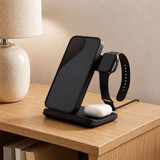 A black 3 in 1 wireless charger for a phone, earbuds, and a smartwatch, placed on a wooden table beside a lamp.