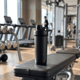 A drink bottle with a double-wall design in black, placed on a gym bench with exercise equipment in the background.