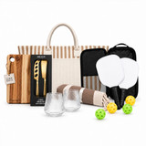 A picnic set featuring a striped tote bag, glasses, a cheese board, utensils, paddles, balls, and a blanket.