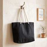 A black recycled beach tote bag with rope handles hangs on a hook against a textured wall.
