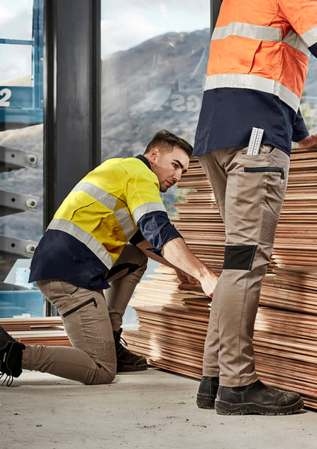 Two workers in high-visibility jackets and stretch cuffed pants, stacked wood planks in an industrial setting.
