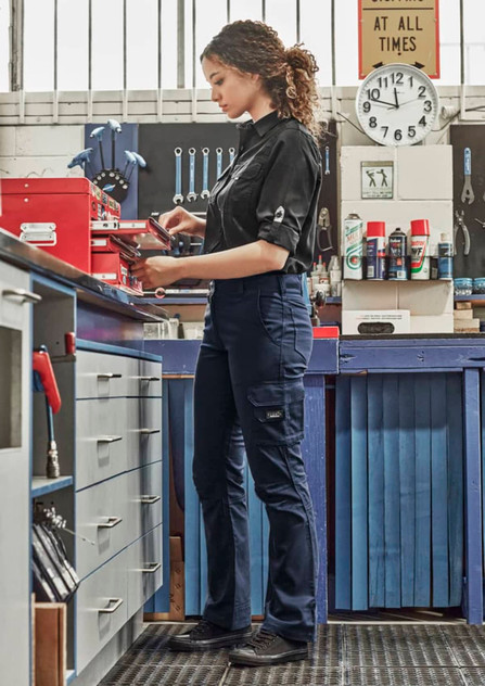 A woman in a black shirt wears navy rugged cargo pants while working in a workshop, surrounded by tools and equipment.