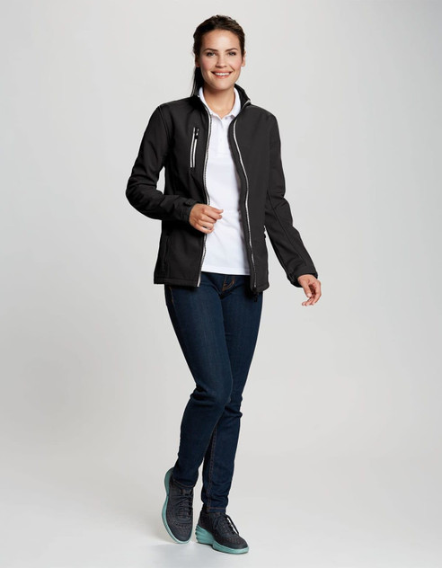 A woman wearing a black softshell jacket over a white top and jeans, featuring a logo on the jacket. A woman wearing a black softshell jacket over a white top and jeans, featuring a logo on the jacket.