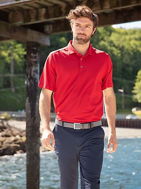 A man wearing a red polo shirt stands by a pier, surrounded by greenery and water. The shirt features a logo.