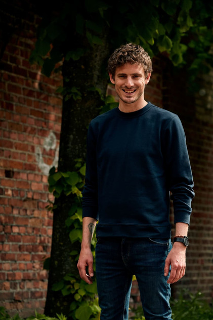 A man wearing a navy crewneck sweatshirt and blue jeans stands against a brick wall with greenery. A man wearing a navy crewneck sweatshirt and blue jeans stands against a brick wall with greenery.