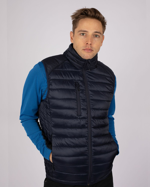 A navy blue men's vest with a zip front and padded design, worn by a model in a blue long-sleeve top. A navy blue men's vest with a zip front and padded design, worn by a model in a blue long-sleeve top.