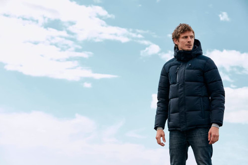 A man stands outdoors wearing a navy puffer jacket against a backdrop of blue skies and clouds. The jacket features a logo. A man stands outdoors wearing a navy puffer jacket against a backdrop of blue skies and clouds. The jacket features a logo.