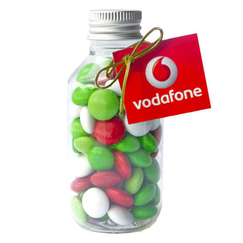A clear glass jar filled with red, green, and white chocolate beans, featuring a swing tag with a logo. A clear glass jar filled with red, green, and white chocolate beans, featuring a swing tag with a logo.