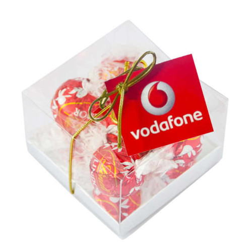 A clear cube filled with five red-wrapped Lindt chocolates, tied with a gold ribbon and featuring a tag. A clear cube filled with five red-wrapped Lindt chocolates, tied with a gold ribbon and featuring a tag.