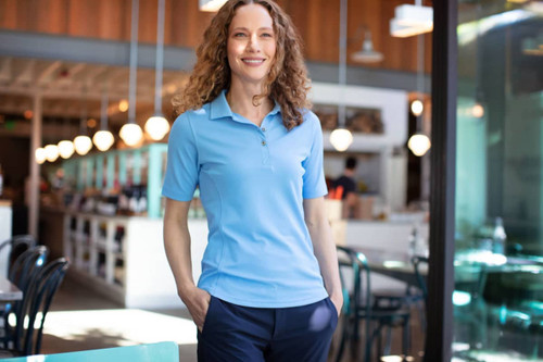 A woman stands in a café wearing a light blue polo shirt paired with dark trousers. The shirt features a logo. A woman stands in a café wearing a light blue polo shirt paired with dark trousers. The shirt features a logo.