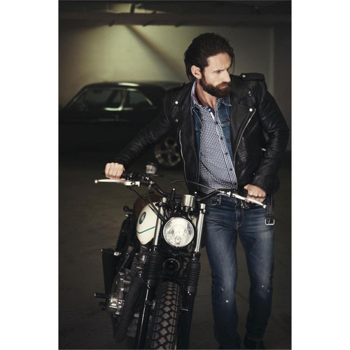 A man in a black leather jacket and checkered shirt stands beside a motorcycle in a dimly lit garage.