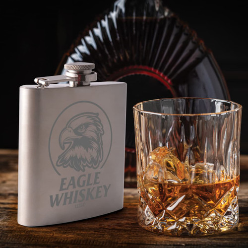 A stainless steel hip flask with an engraved design beside a glass of whiskey containing ice. A stainless steel hip flask with an engraved design beside a glass of whiskey containing ice.