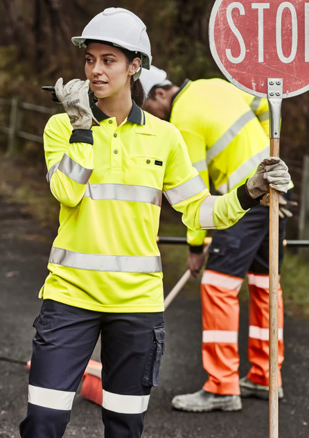 A woman in a high-visibility yellow polo shirt with reflective tape, wearing a hard hat and holding a sign.
