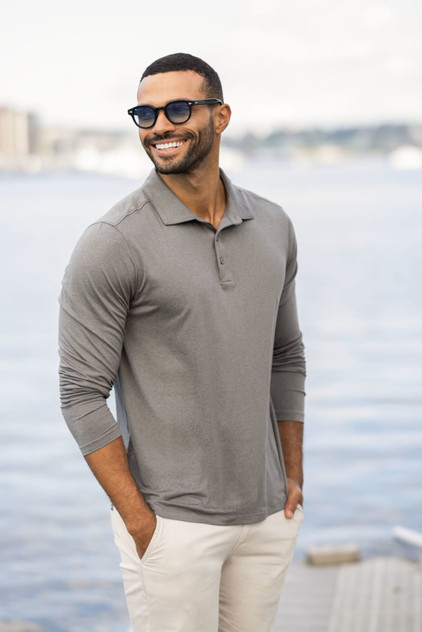A man wearing a grey long sleeve polo shirt stands by the water, smiling, with sunglasses and light-coloured pants.
