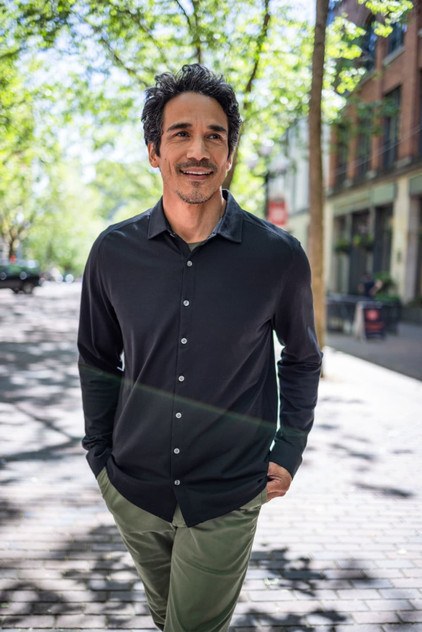 A long-sleeved, button-up shirt in black, modelled by a man outdoors in a tree-lined street.