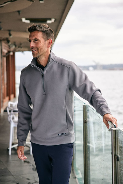 A grey half zip jacket worn by a man standing near a railing by the water, featuring a logo.