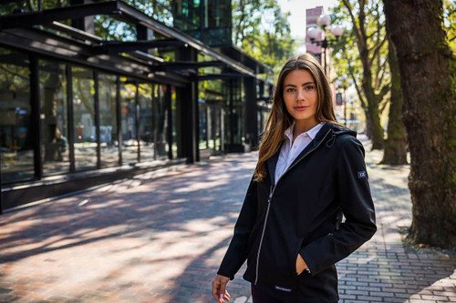 A woman stands outside wearing a black softshell jacket. The jacket features a hood and has a logo. A woman stands outside wearing a black softshell jacket. The jacket features a hood and has a logo.