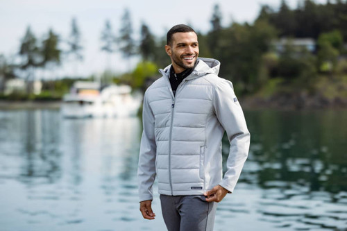 A man wearing a light grey hybrid jacket with a hood, smiling by the water, surrounded by trees and boats.