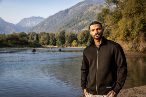 A man stands by a river wearing a black full zip jacket against a scenic mountain backdrop. The jacket features a logo. A man stands by a river wearing a black full zip jacket against a scenic mountain backdrop. The jacket features a logo.