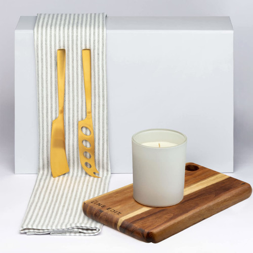 A wooden cheese board with a handle, a gold cheese knife, a spatula, and a white candle on a grey background.