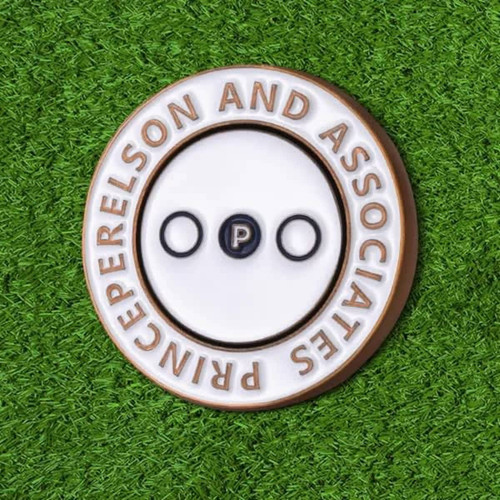 A round, soft enamel magnetic ball marker in gold and white with a central black dot, set against a grassy background.