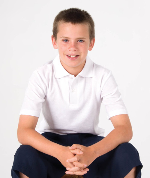 A white kid's pique polo shirt worn by a boy sitting, with dark shorts and a neutral background. The shirt has a logo.