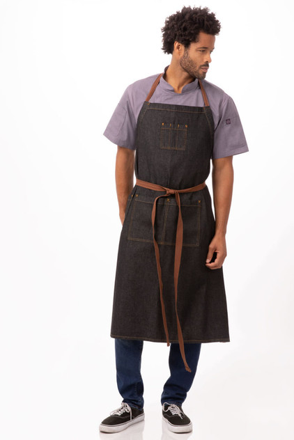 A dark denim apron with brown straps, featuring pockets and worn by a model standing gracefully.