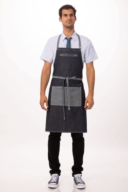 A man wearing a dark denim bib apron with grey accents and pockets, paired with a light shirt and tie.