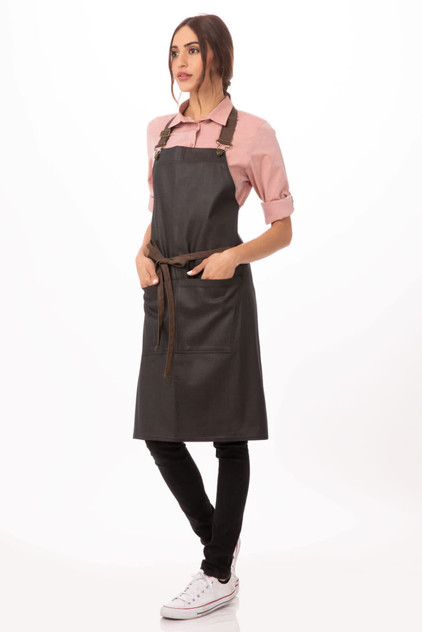 A woman wearing a black Boulder Bib Apron with brown straps, paired with a pink shirt and black pants.