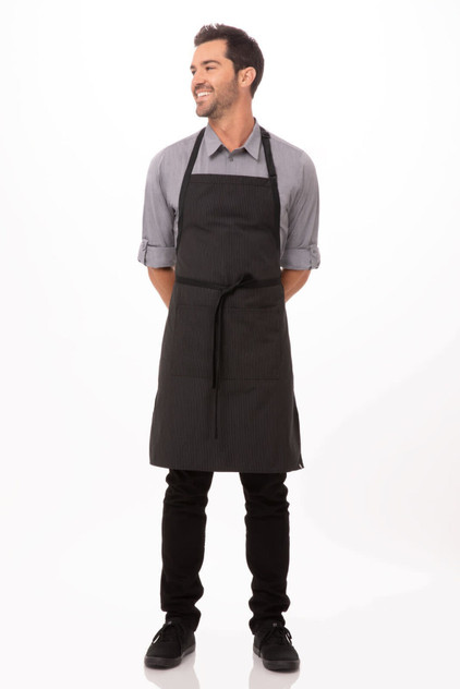A black bib apron with contrasting ties worn by a smiling man, standing upright with hands behind his back.
