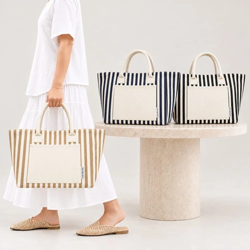 A striped tote bag in beige and cream, with additional navy and white options, displayed on a stone table.