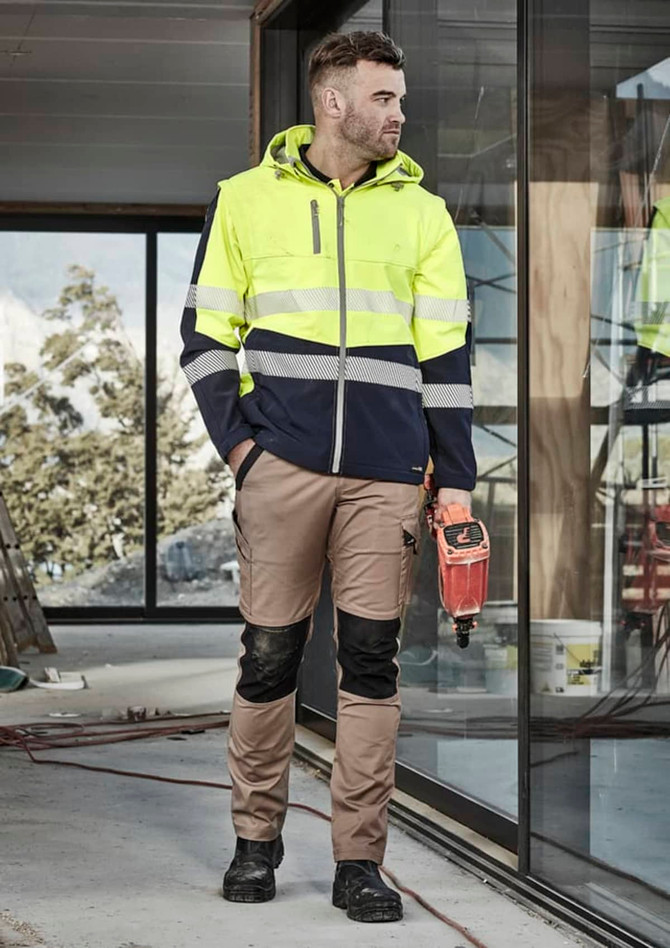A man wearing a high-visibility yellow and navy work jacket and khaki work pants, standing in a construction setting.