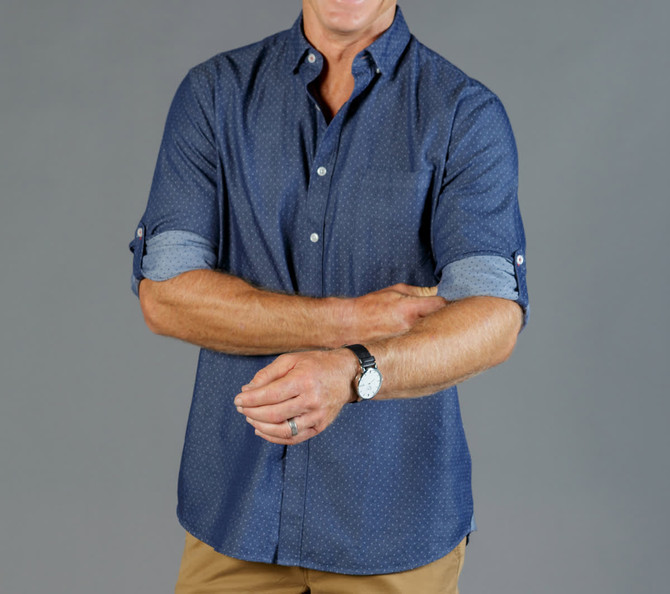 A button-up shirt in navy chambray with rolled sleeves and a subtle pattern. It features a logo on the chest.