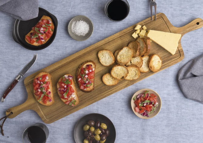 A wooden tapas serving board features bruschetta, crackers, cheese, olives, and drinks on a textured blue tablecloth.
