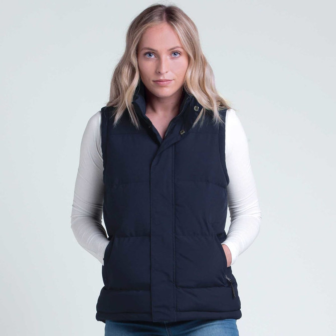 A woman wearing a navy puffa vest over a long-sleeve white top, with a branding logo visible.