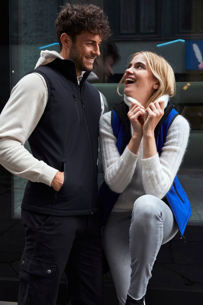 A black softshell vest is worn by a man, while a woman smiles in a blue softshell vest, both against a modern backdrop.