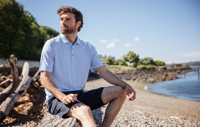 A man wearing a light blue botanical print polo shirt sits on a log by the water, set against a scenic background. A man wearing a light blue botanical print polo shirt sits on a log by the water, set against a scenic background.