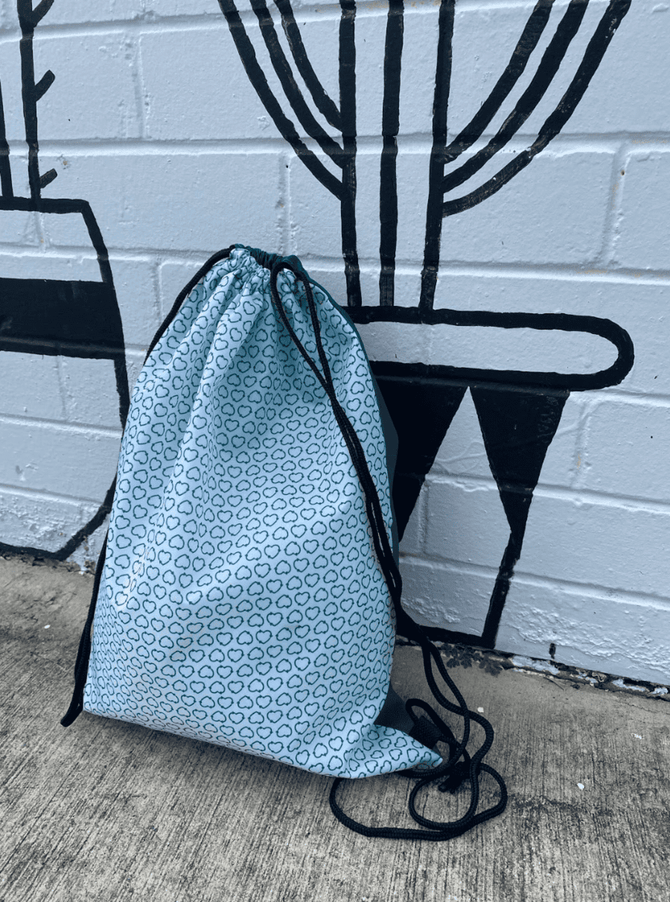 A light blue drawstring backpack with a circular pattern, featuring black cords, against a brick wall with a mural.