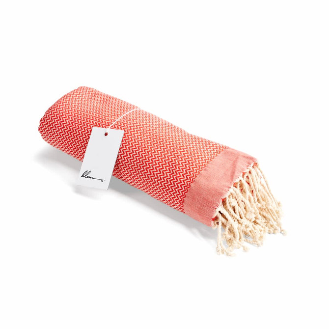 A rolled coral and red beach towel with fringed edges and a tag attached, featuring a chevron pattern.
